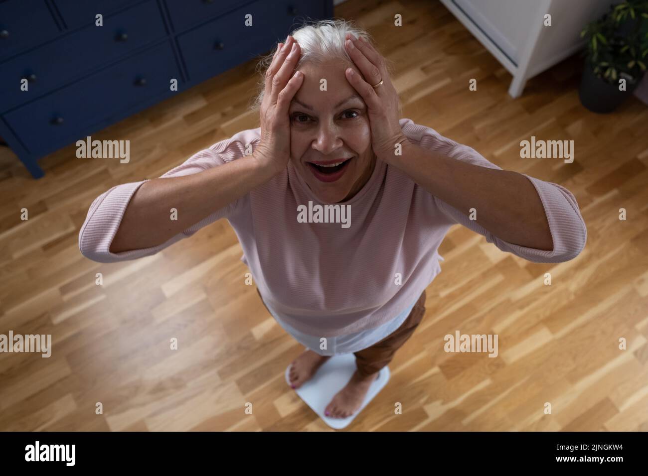 Senior worried woman stepping on floor scales, holding hands on head being shocked. Weight control Stock Photo
