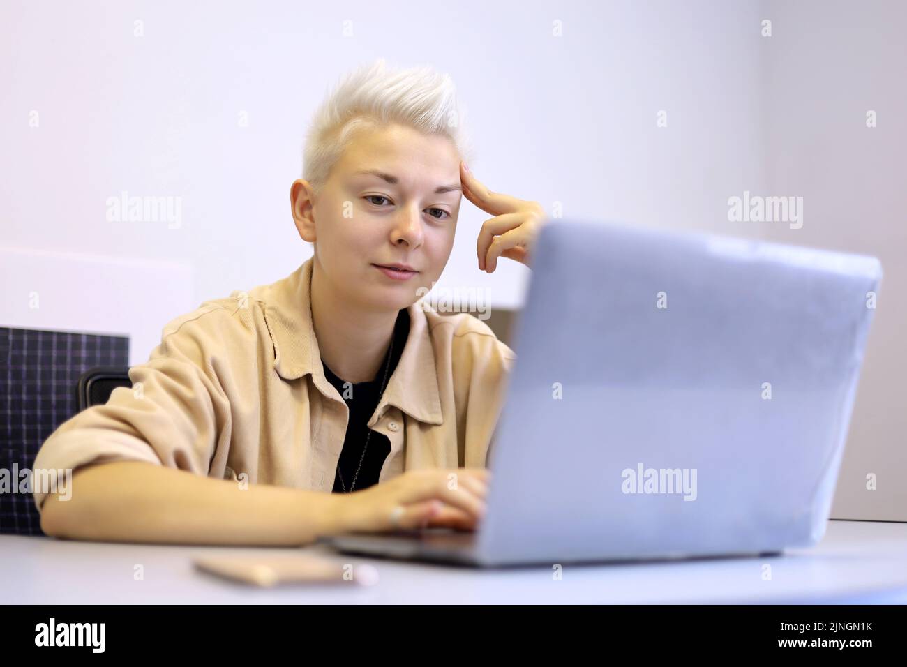 Smiling girl with short blonde hair sitting at office table with laptop. Tomboy lifestyle, concept of inspiration at work and creativity Stock Photo