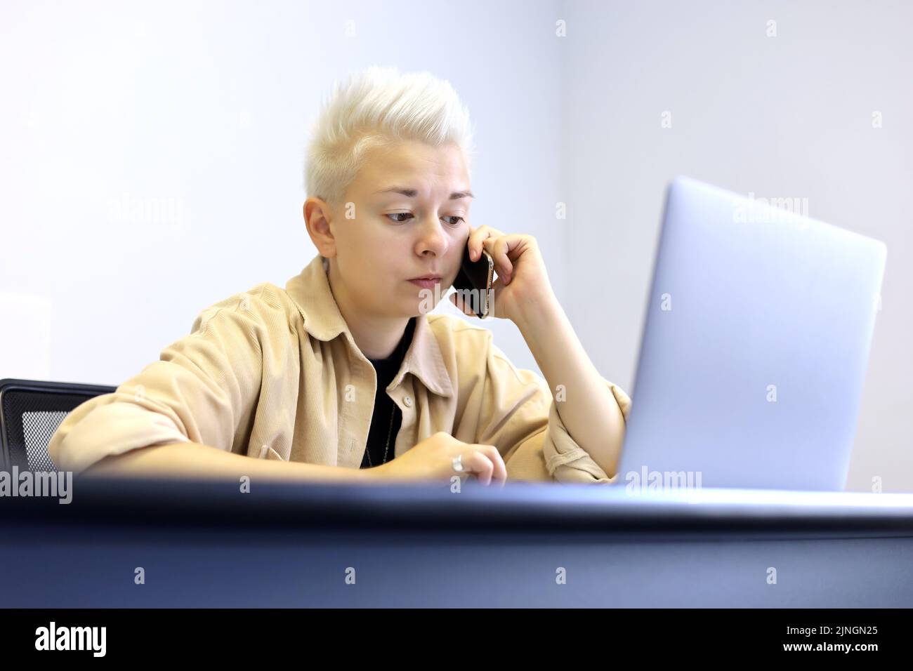 Girl with short blonde hair sitting at laptop and talking on mobile phone. Tomboy lifestyle, concept of office work, business assistant Stock Photo