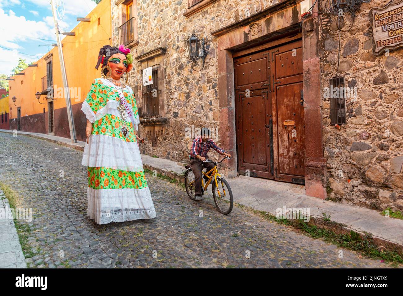 A bicyclist passes a mojiganga, or giant puppet made from papier mâché as it walks through the historic city center of San Miguel de Allende, Mexico. Stock Photo