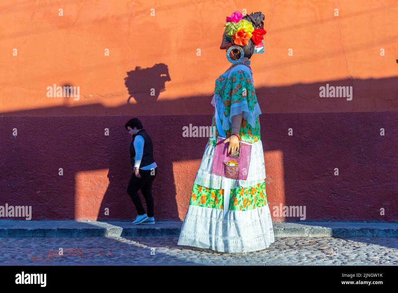 A woman passes a mojiganga, or giant puppet made from papier mâché as it walks through the historic city center of San Miguel de Allende, Mexico. Stock Photo