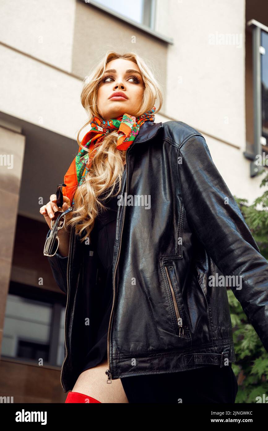Blonde parisian girl with perfect makeup in stylish colorful headscarf, vintage leather jacket holding in hand sunglasses walking down street. Paris Stock Photo