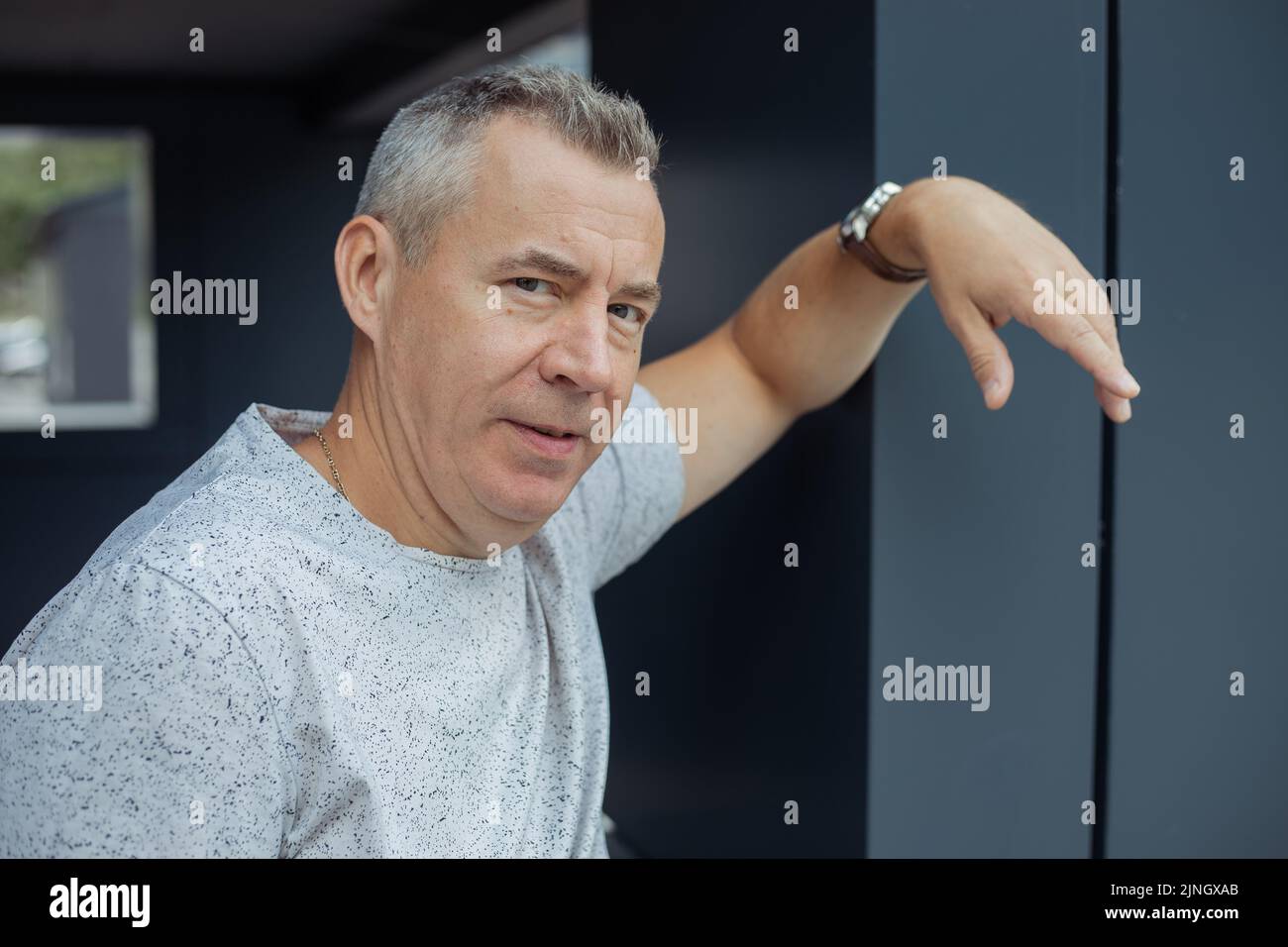 Portrait closeup of attractive aged gray haired man leaning arm on gray construction of modern office building, selective focus. Urban lifestyle Stock Photo