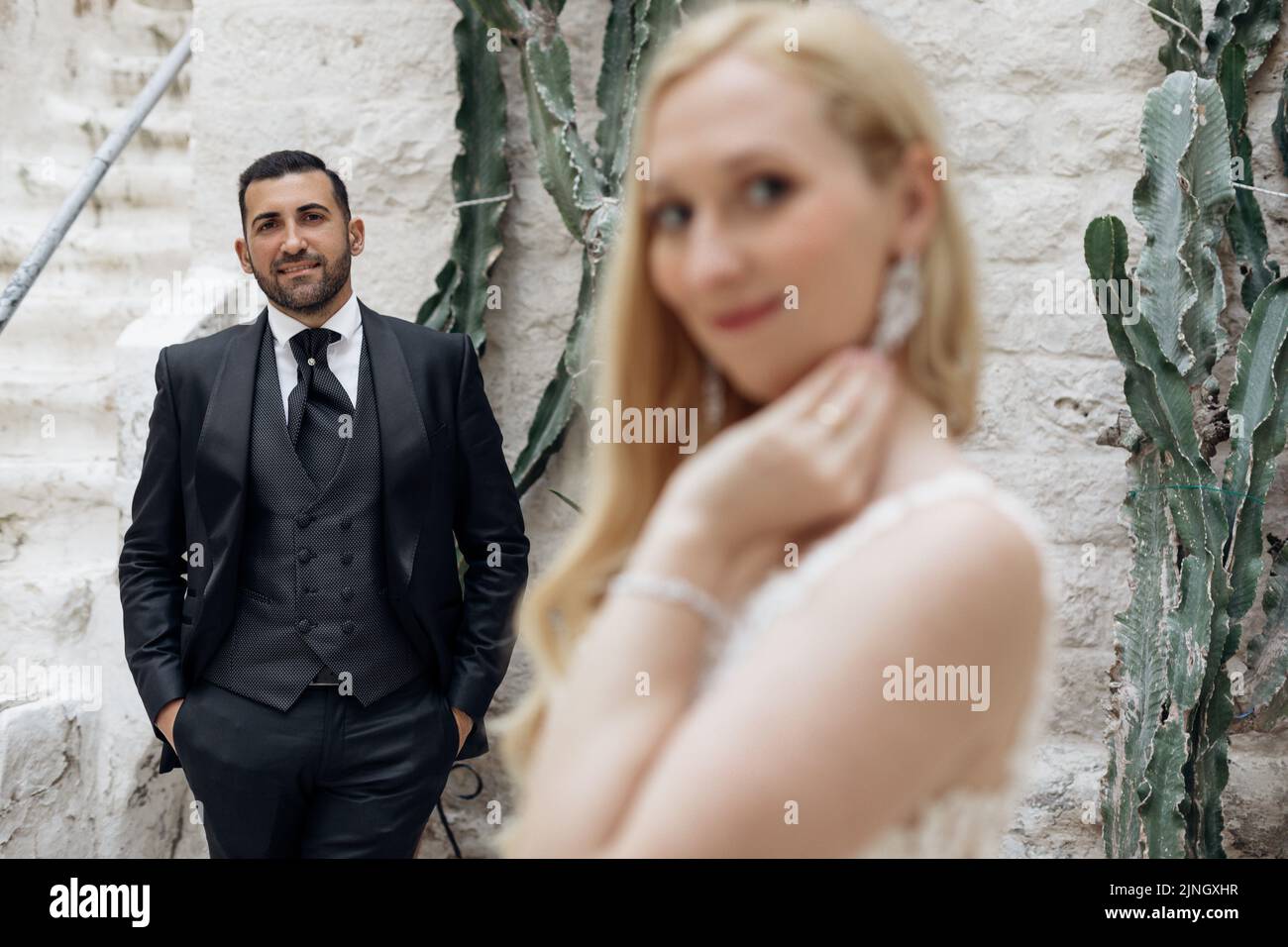 Portrait of wedding couple standing near white brick wall. Groom in suit admiring bride, jamming hands into pockets.  Stock Photo