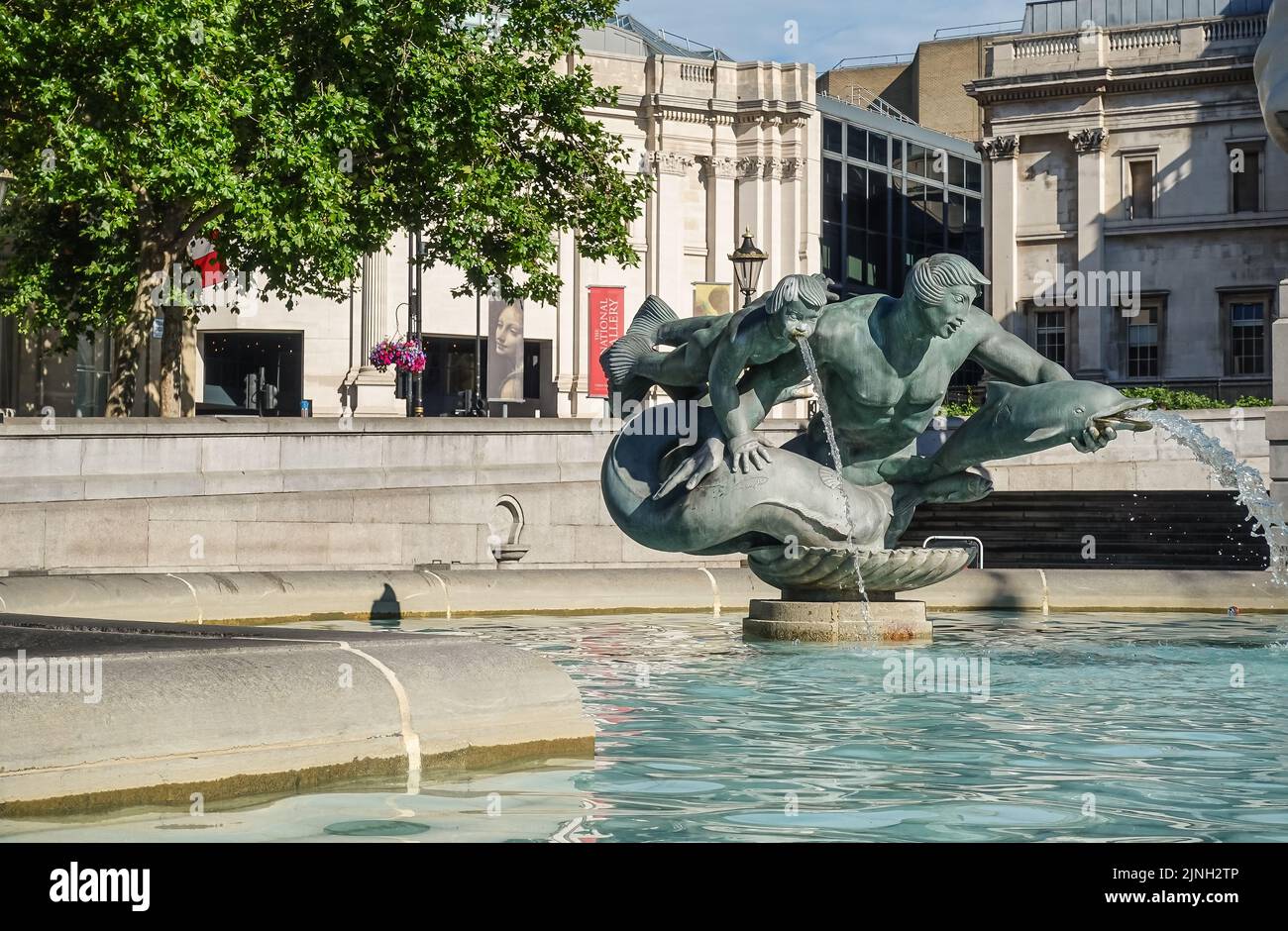 London, UK- July 4, 2022: Trafalgar Square. Mermain statue in western pool-fountain with greenf foliage and part of National Gallery facade in back. Stock Photo