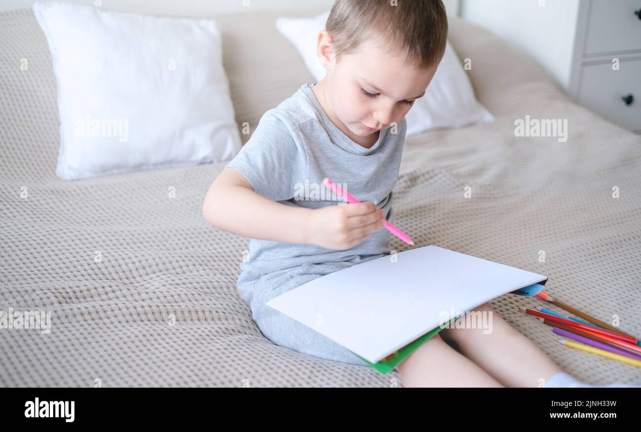 A small caucasian boy in a gray T-shirt and shorts on the bed draws with colored pencils in a sketch Stock Photo