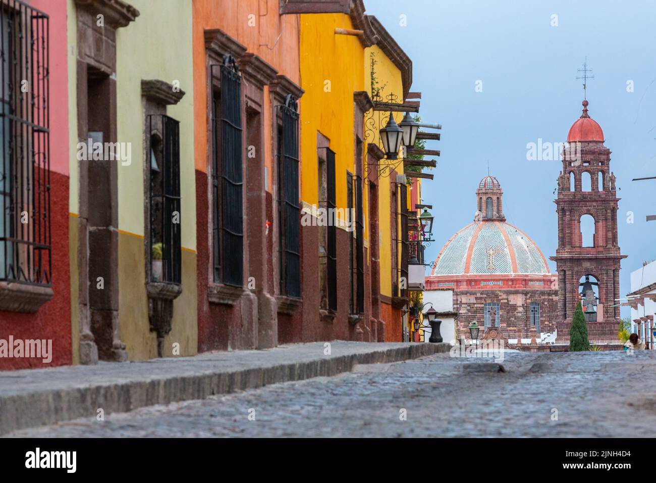The neoclassical style dome and bell tower of Iglesia de San Francisco peaks over the cobblestone road on Calle Recreo in the historic city center of San Miguel de Allende, Mexico. Stock Photo