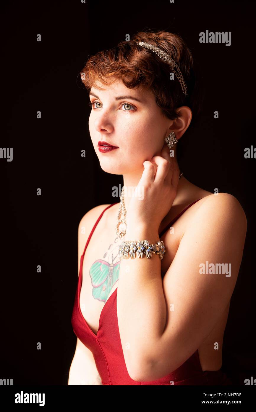 Self-portrait. Window-lit portrait of a woman with short hair wearing a red gown and vintage jewelry. Red lips, hazel eyes, brunette Stock Photo
