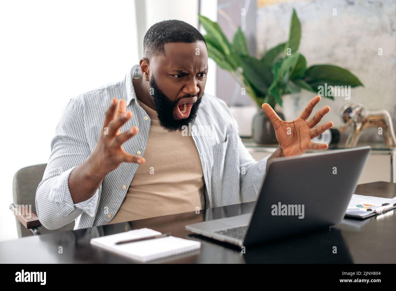Irritated angry african american man, company worker, freelancer, ceo company, sitting at a desk in modern office, yelling at the laptop, dissatisfied with the result, annoyed emotional state Stock Photo