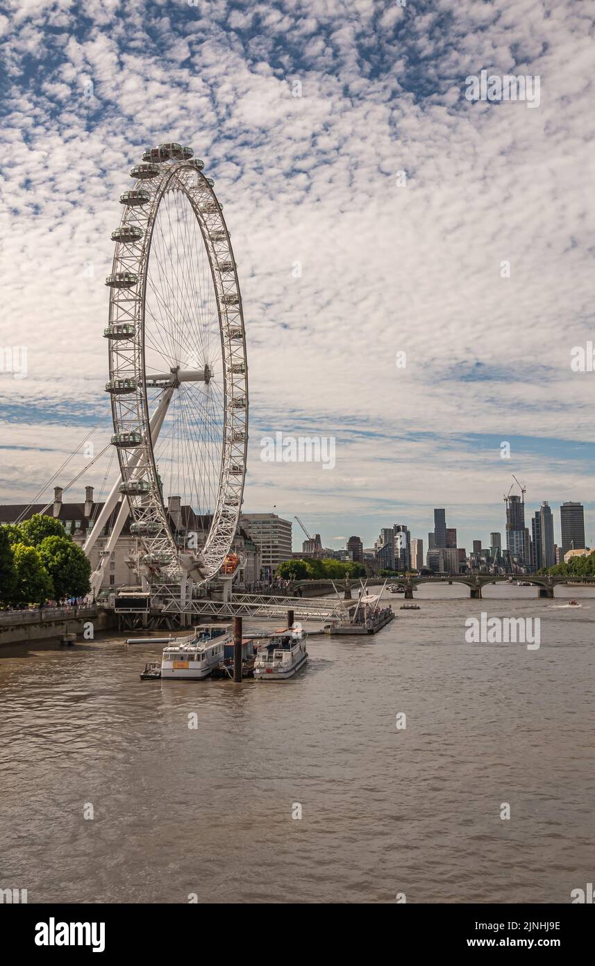 London, UK - July 4, 2022: Closeup of White London Eye sidewats against blue cloudscape. Brown water Thames River in front. Cityscape of multi-story b Stock Photo