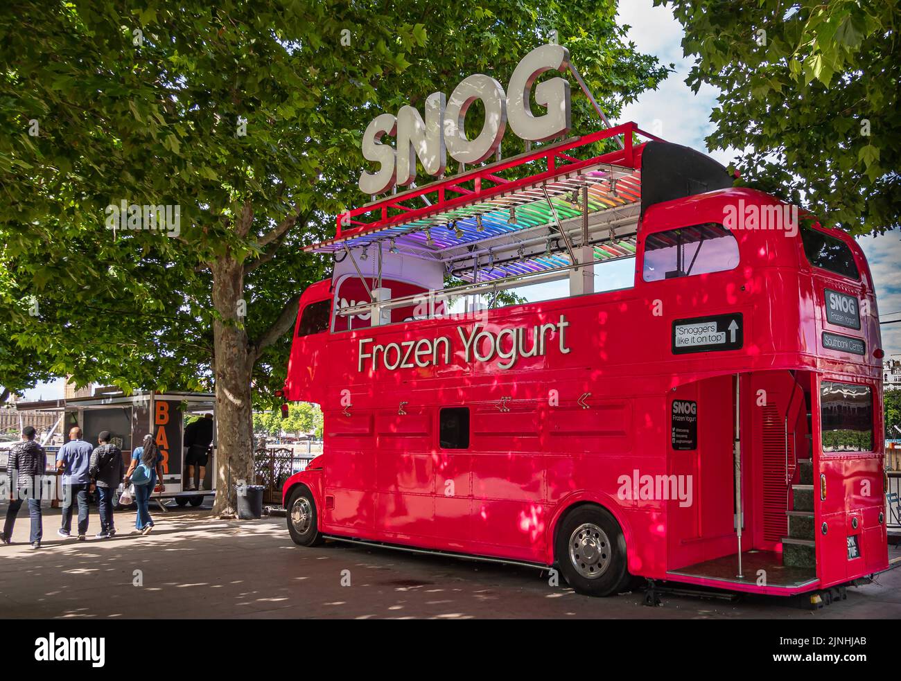 London, UK - July 4, 2022: Red SNOG Frozen Yogurt bus parked in Jubilee Park under green foliage. People present. Stock Photo