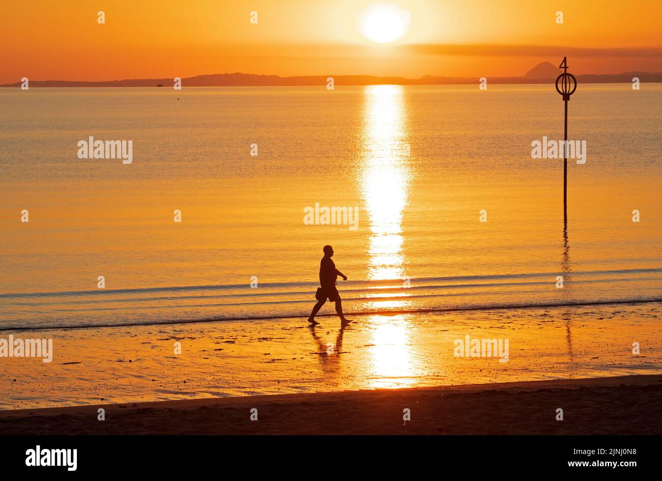 Portobello, Edinburgh, Scotland, UK. 12th August 2022. Stunning sunrise to end a very hot week of above average temperatures for Scotland, temperature this morning at dawn 13 degrees centigrade for those out for exercise at the Firth of Forth seaside with haar expected later on the east coast.. Pictured: A man walks along the shore at dawn. Credit: Arch White/alamy live news. Stock Photo