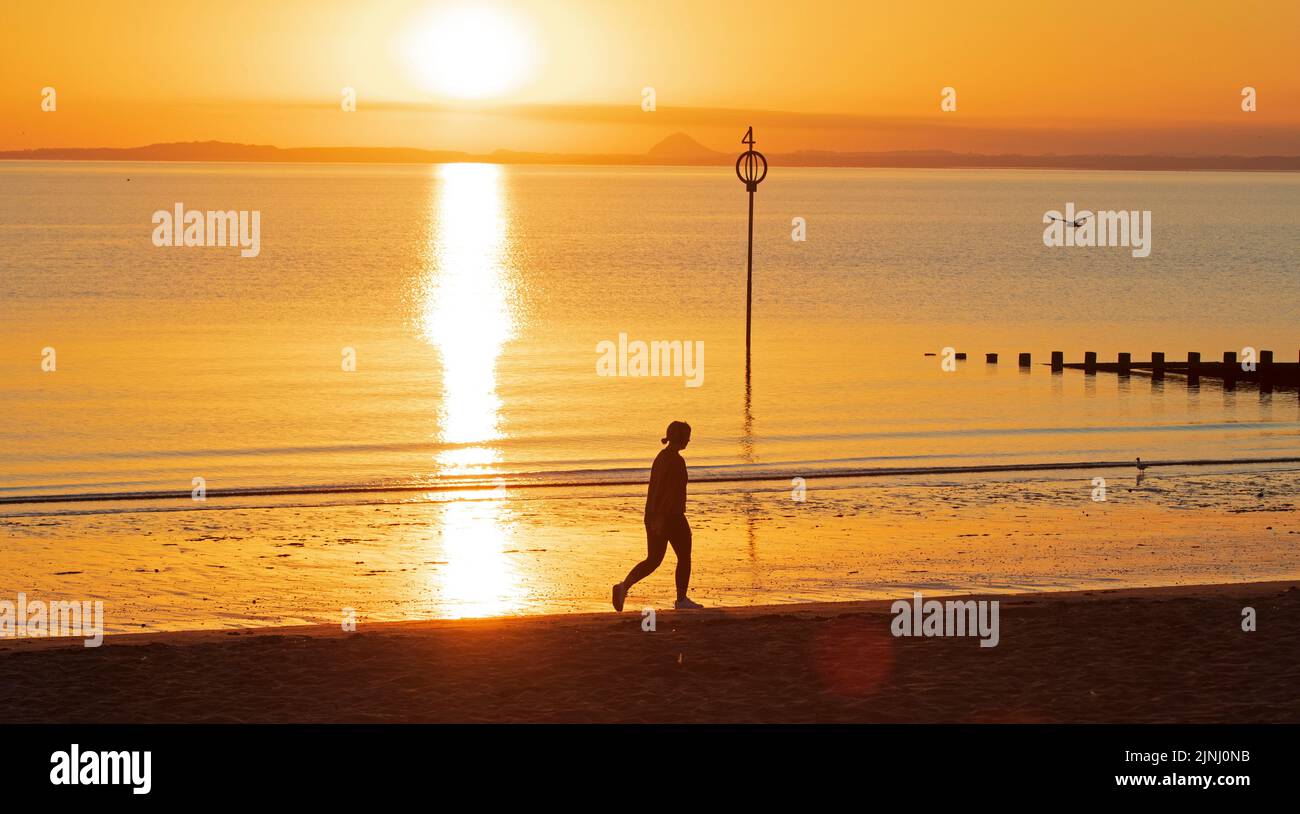 Portobello, Edinburgh, Scotland, UK. 12th August 2022. Stunning sunrise to end a very hot week of above average temperatures for Scotland, temperature this morning at dawn 13 degrees centigrade for those out for exercise at the Firth of Forth seasid , with haar expected later on the east coast.. Pictured: A woman walks along the shore at dawn. Credit: Arch White/alamy live news. Stock Photo