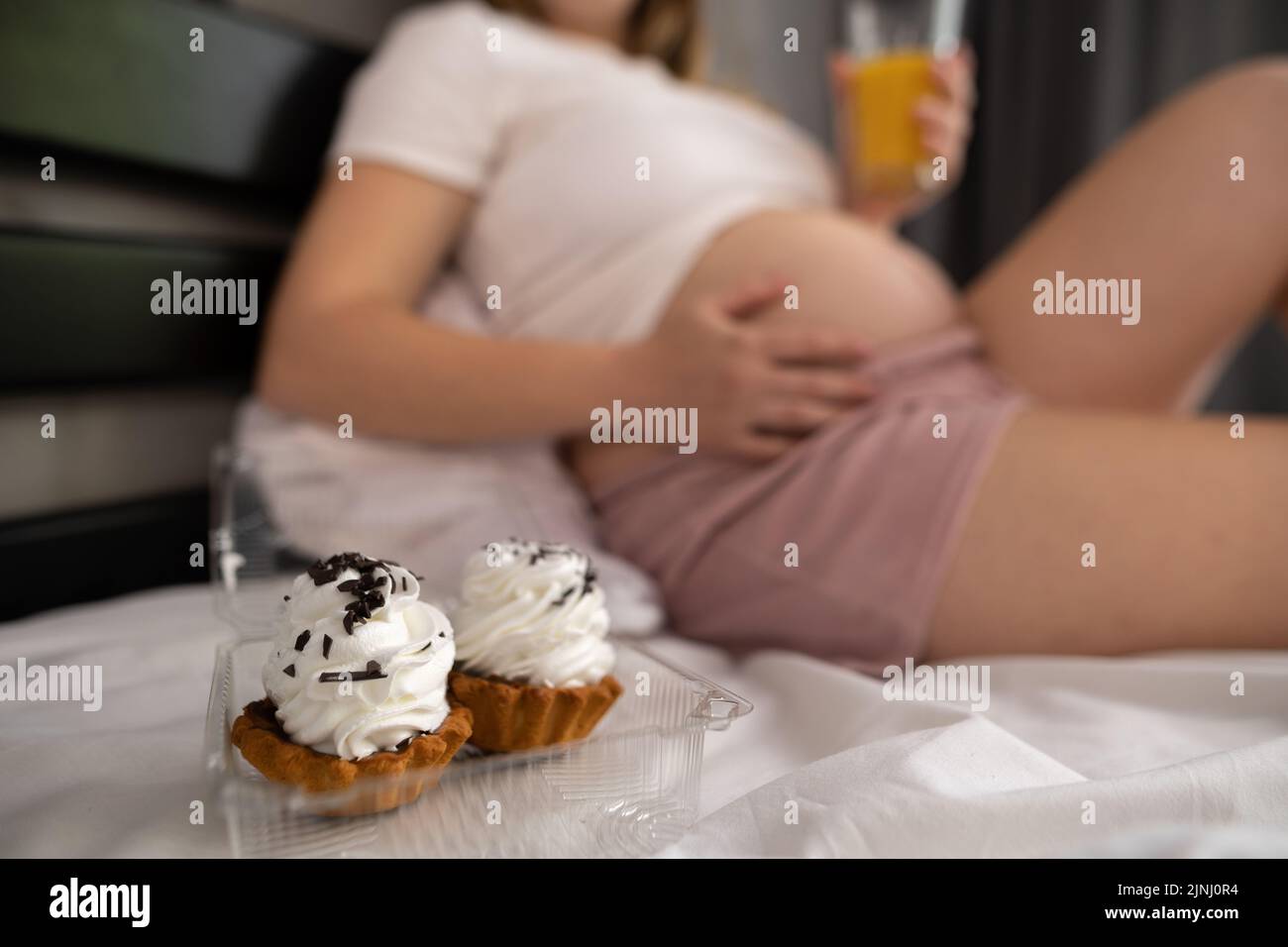 Close up of pregnant woman staying in bed to eat junk cakes. Pregnancy and unhealthy eating concept Stock Photo