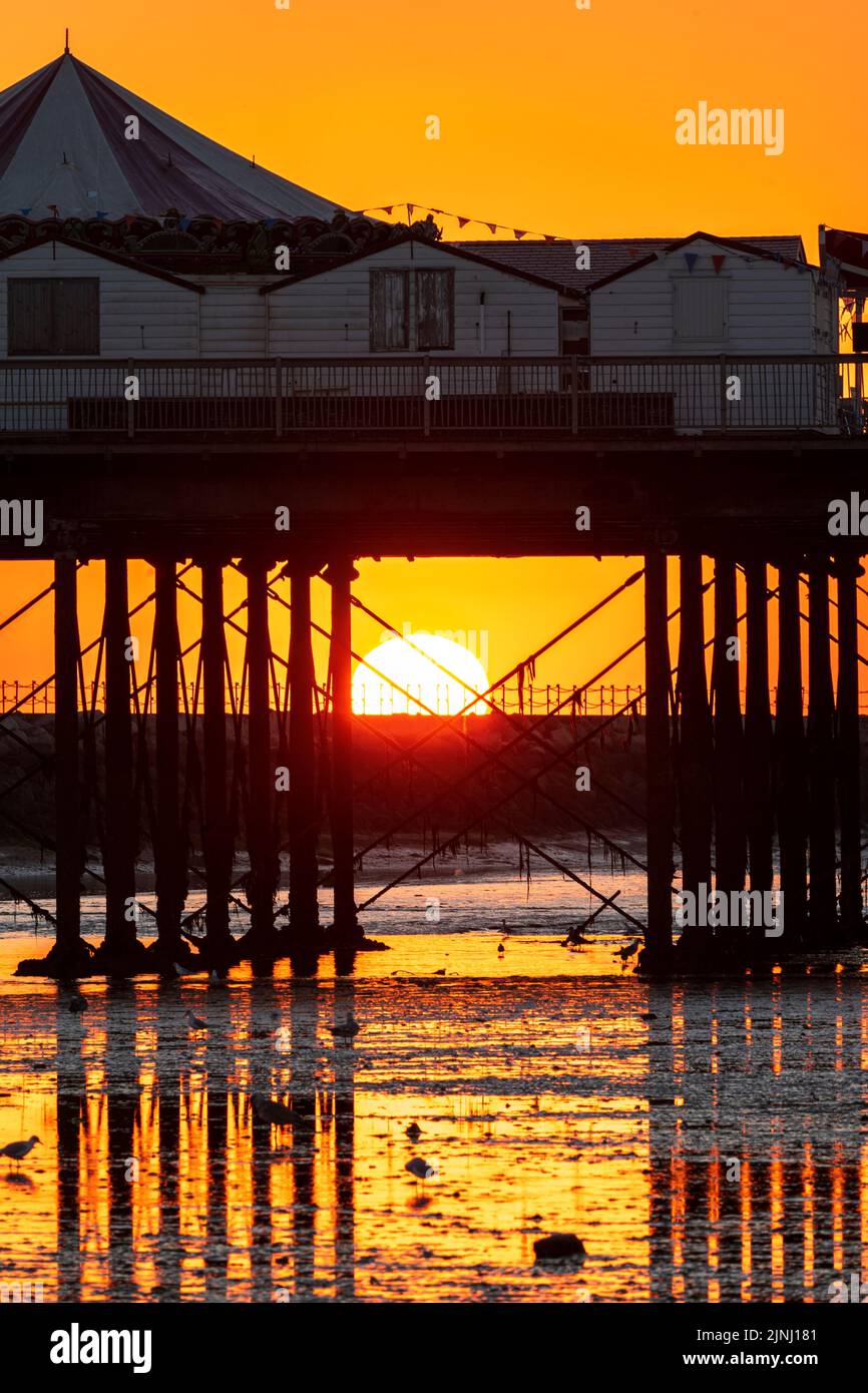 Close up of the sunrise seen through the pillars supporting the pier at low tide at the Kent resort town of Herne Bay on a summer's morning. Stock Photo