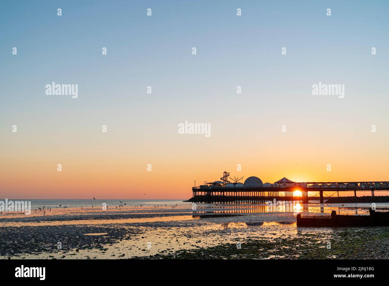 Sunrise behind the pier at the Kent resort town of Herne Bay on a summer's morning. View along the beach at low tide to the pier with the sun rising directly behind it. Stock Photo