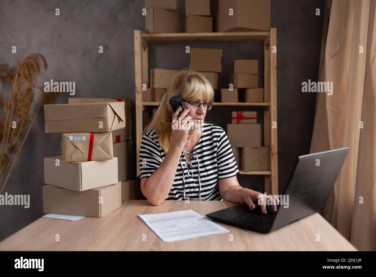 Older female dropshipping business owner using laptop talking on phone in warehouse. Mature woman seller merchant checking online order, e-commerce Stock Photo