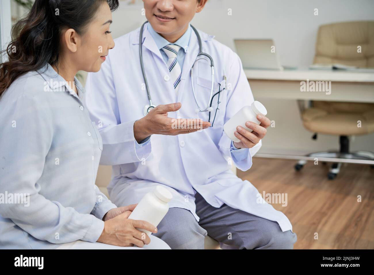 Profile view of senior Asian patient listening to her doctor with interest while he giving her instructions how to take pills correctly, interior of modern office on background Stock Photo