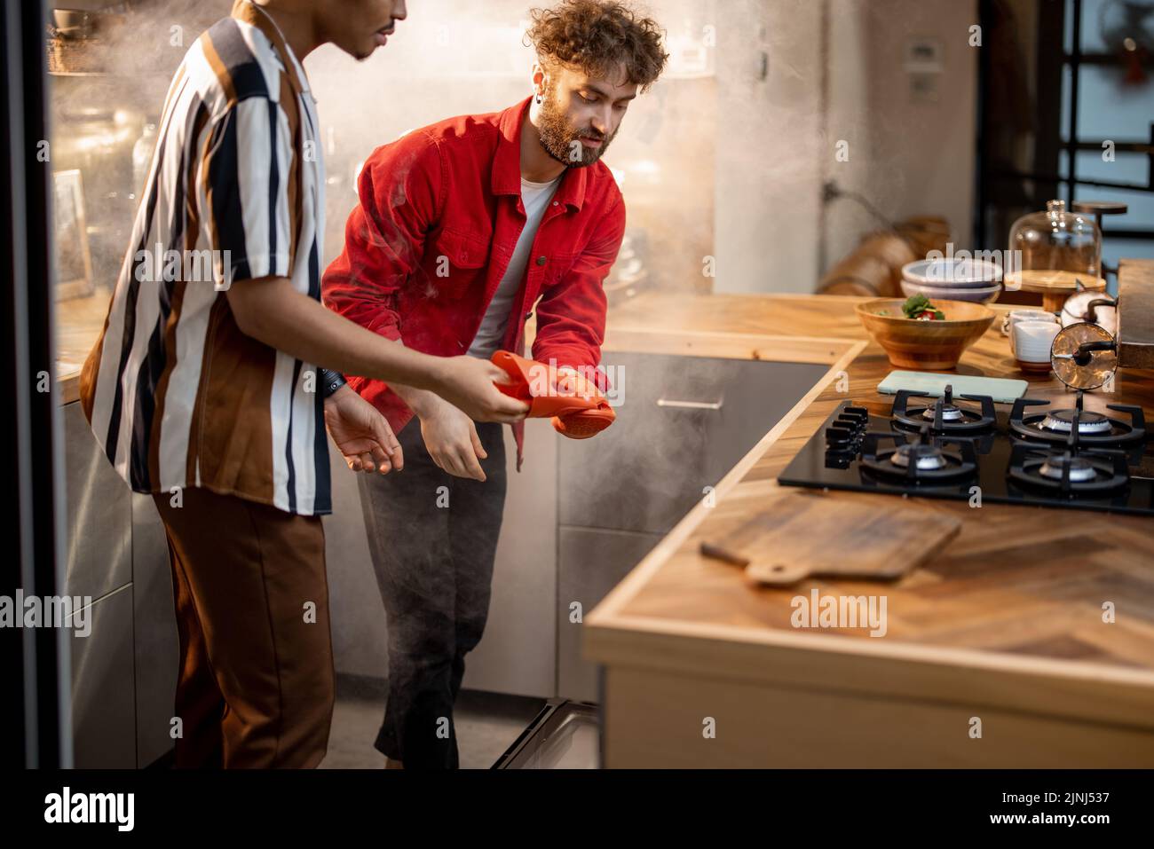 Two guys open the oven while cooking delicious food on kitchen. Steam coming out of the oven. Concept of two gay couple cooking at home Stock Photo