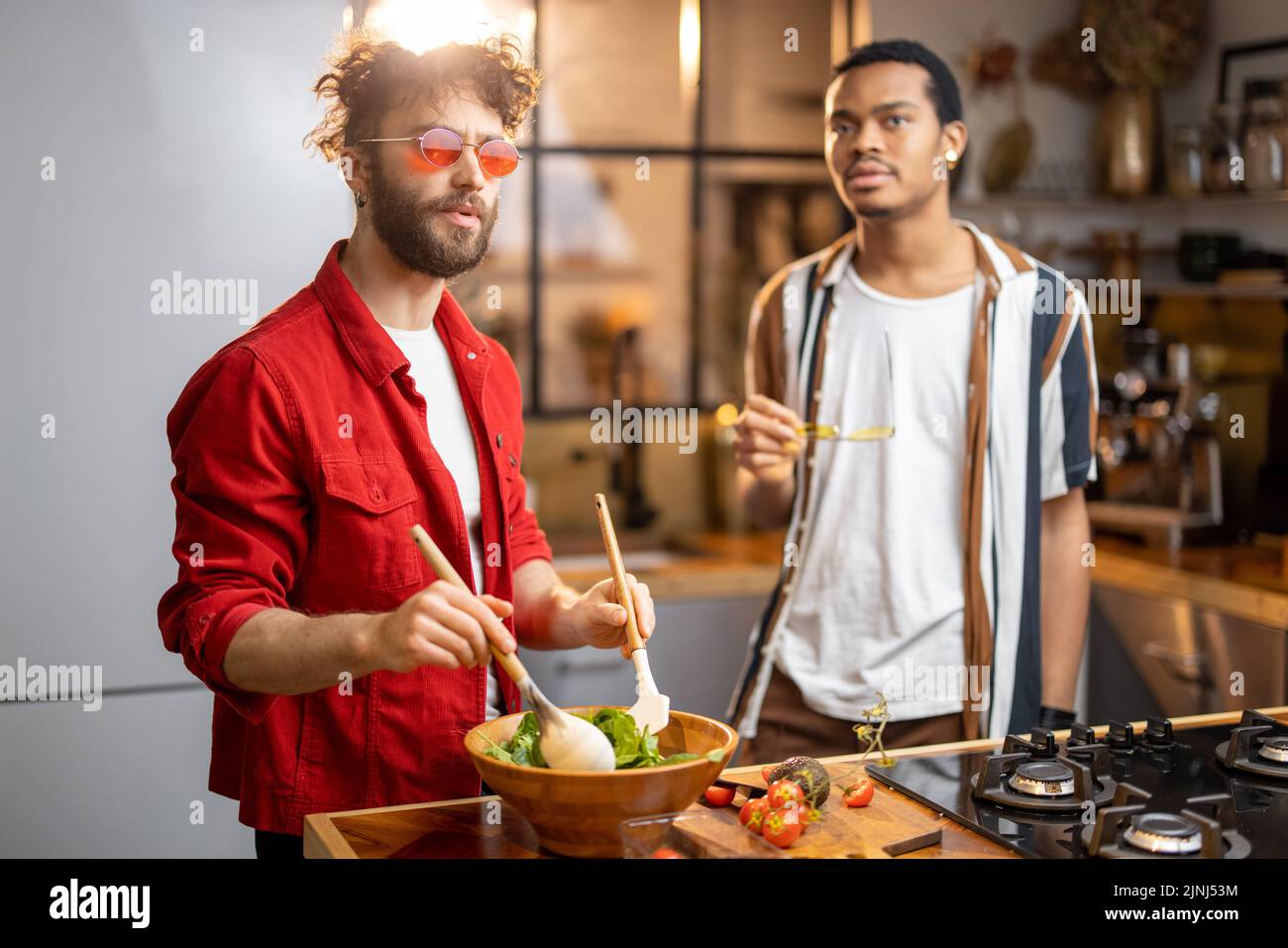 Two brightly dressed guys having fun while making salad together on kitchen. Concept of gay couples and everyday life at home. Caucasian and hispanic Stock Photo