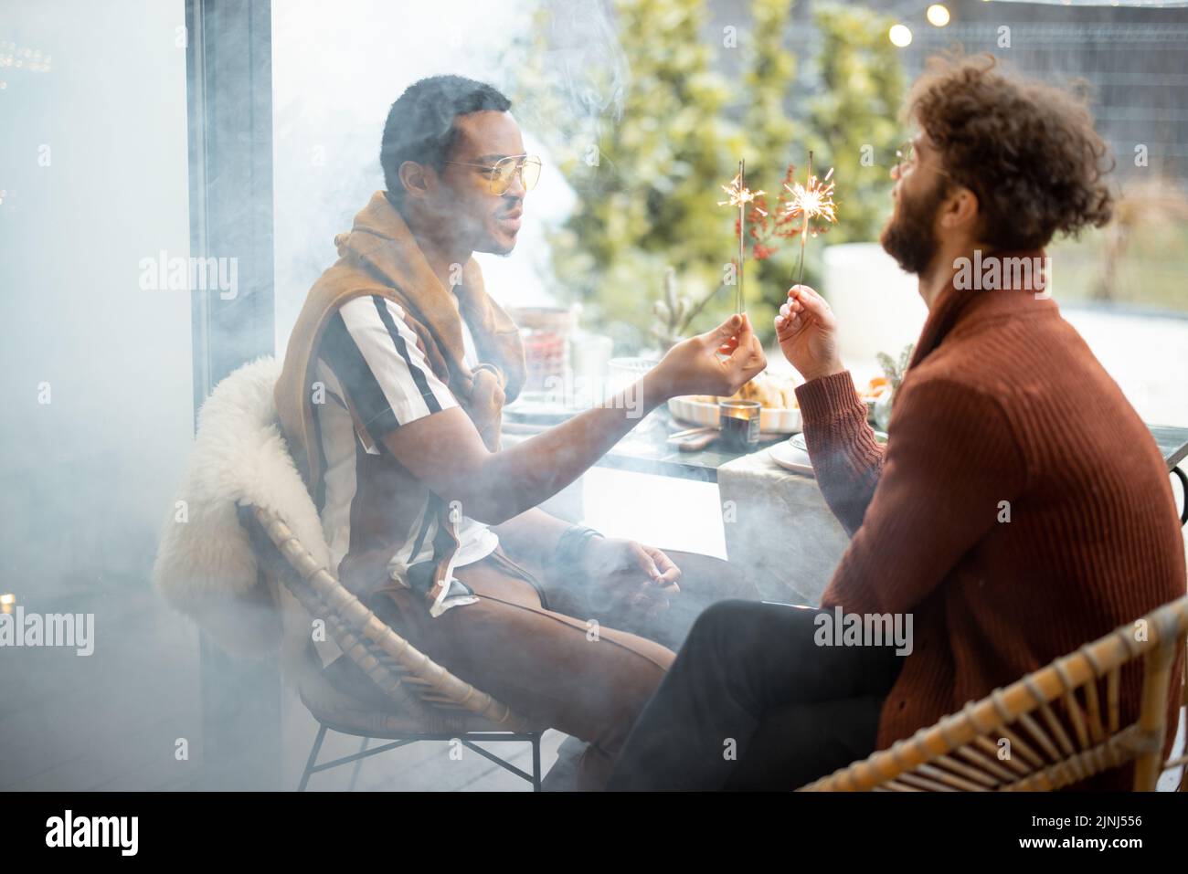 Two male friends having fun burning sparklers, during a festive dinner at backyard. Caucasian and hispanic man celebrating together. Idea of gay coupl Stock Photo