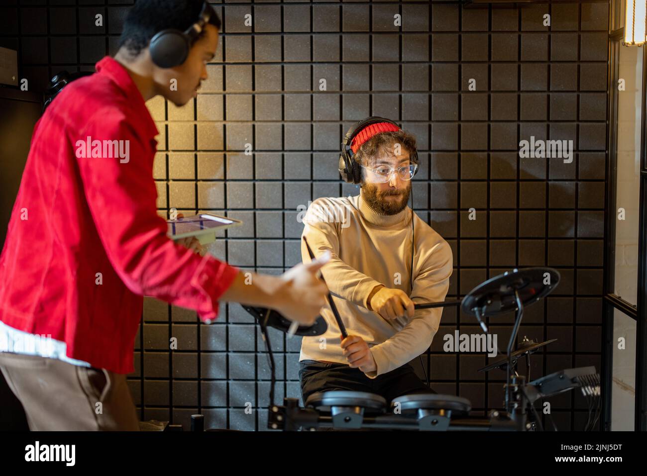 Two stylish men playing electric drums, composing electronic music at small recording studio. Small band creating or practicing music in a home studio Stock Photo