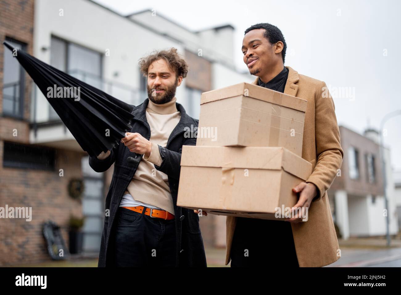 Stylish gay couple walking together with parcels on some residential street on rainy weather. Caucasian and hispanic man with umbrella wearing coats. Stock Photo