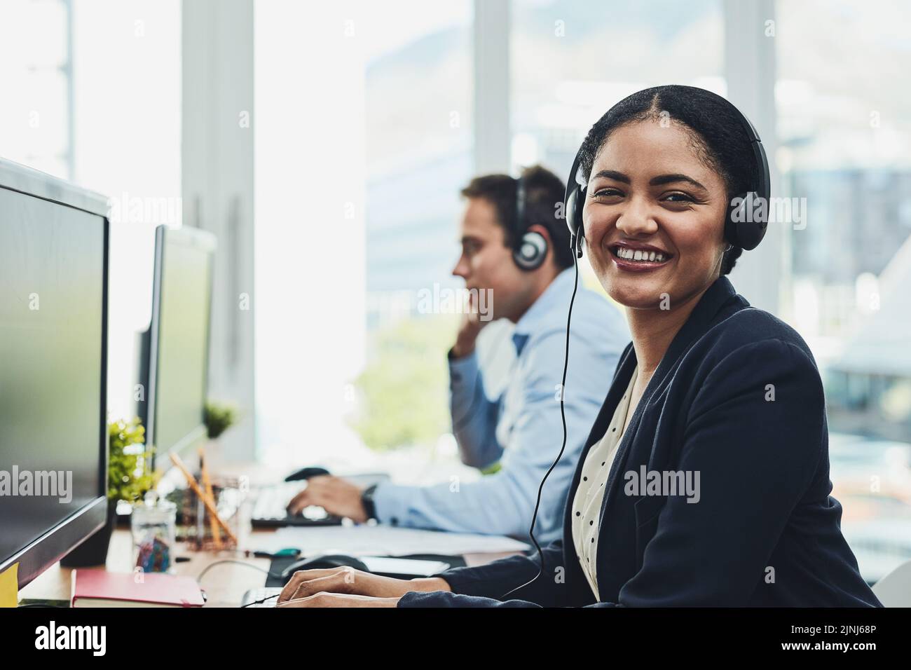 Portrait of happy call center agent working in a busy office, assisting clients and providing good customer service. Young, smiling and cheerful Stock Photo