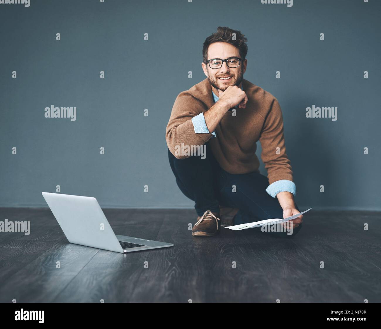 Happy business man working on laptop, doing tax paperwork and going through finance document while sitting on floor in an office at work. Portrait of Stock Photo