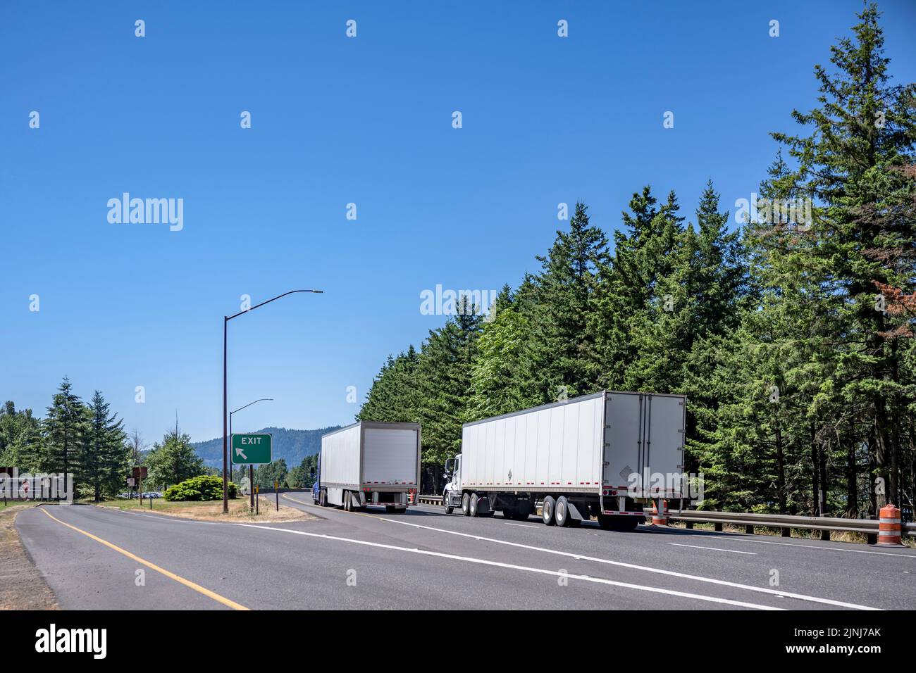 Convoy of tow different industrial freight big rigs semi trucks with dry van semi trailers transporting cargo running on the turning highway road with Stock Photo