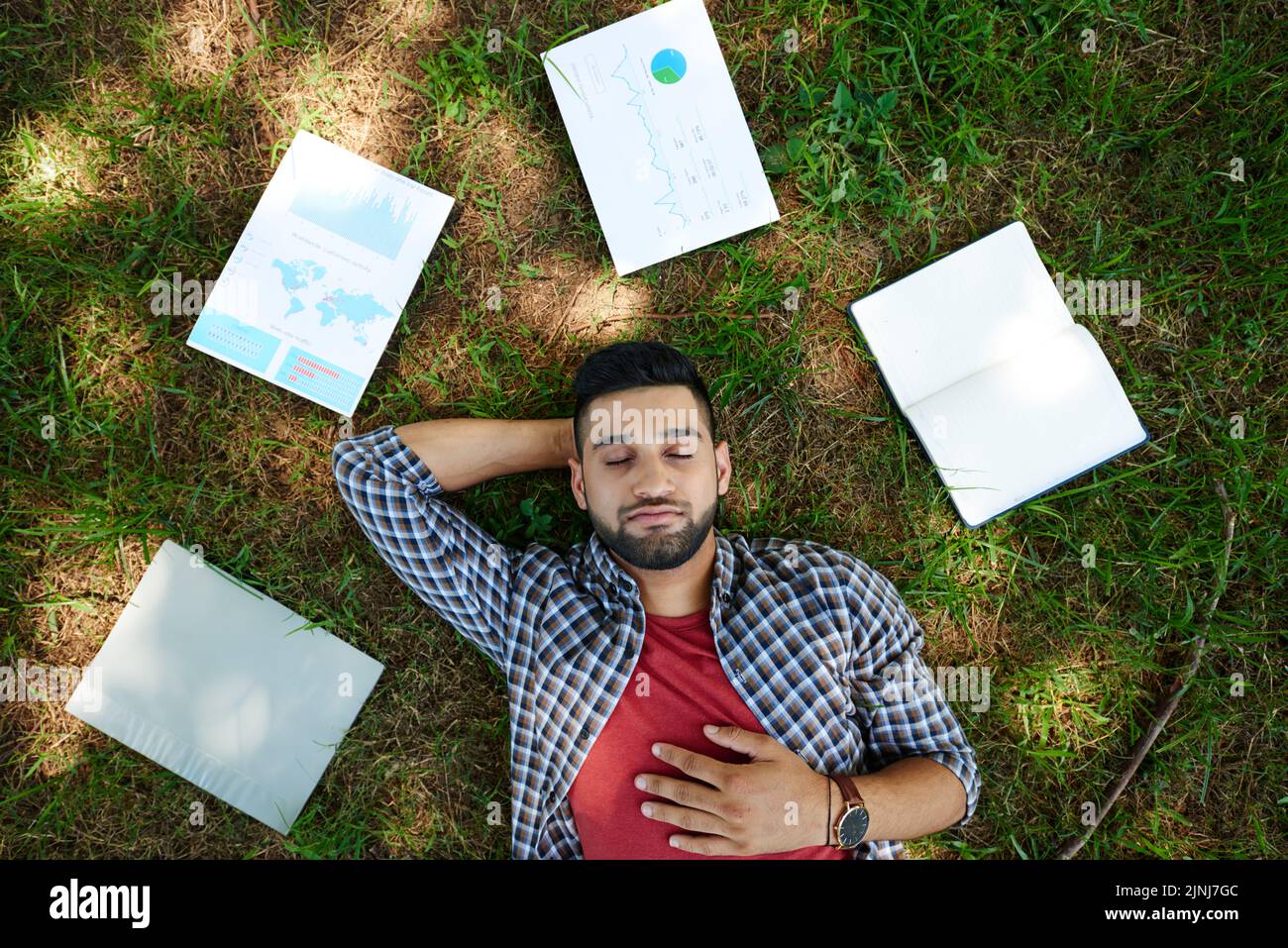 Handsome bearded manager lying on green lawn with closed eyes while taking break from work, documents and notepad illuminated with sunbeams Stock Photo