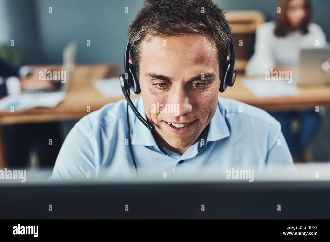 Closeup of a young male call center agent consulting and giving support while working in a busy office. Face of a man and helpdesk worker or phone Stock Photo