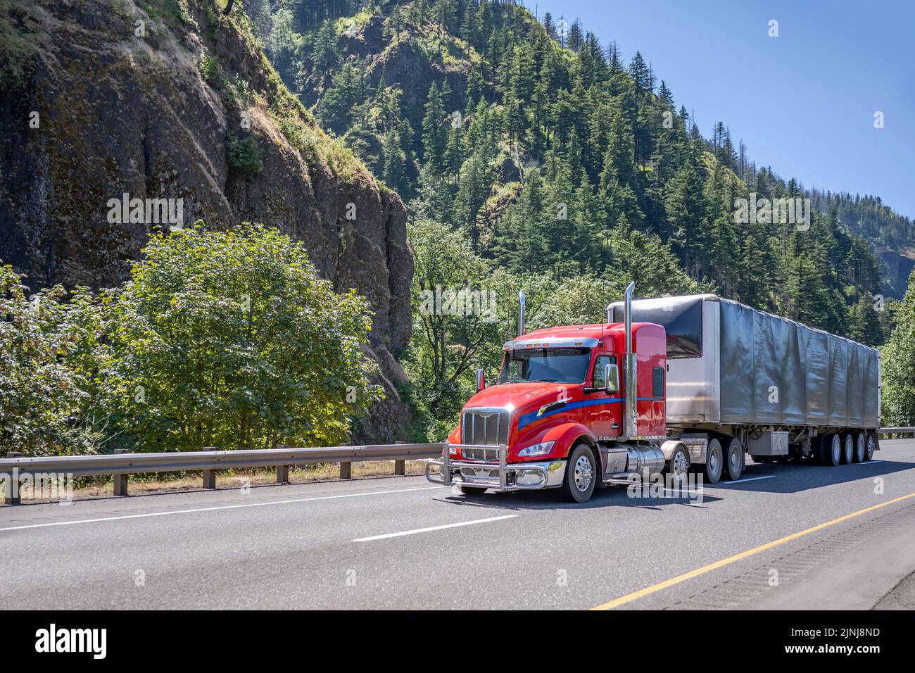 Red industrial low cab profile big rig semi truck transporting commercial cargo in covered framed dry van semi trailer moving on the wide highway road Stock Photo