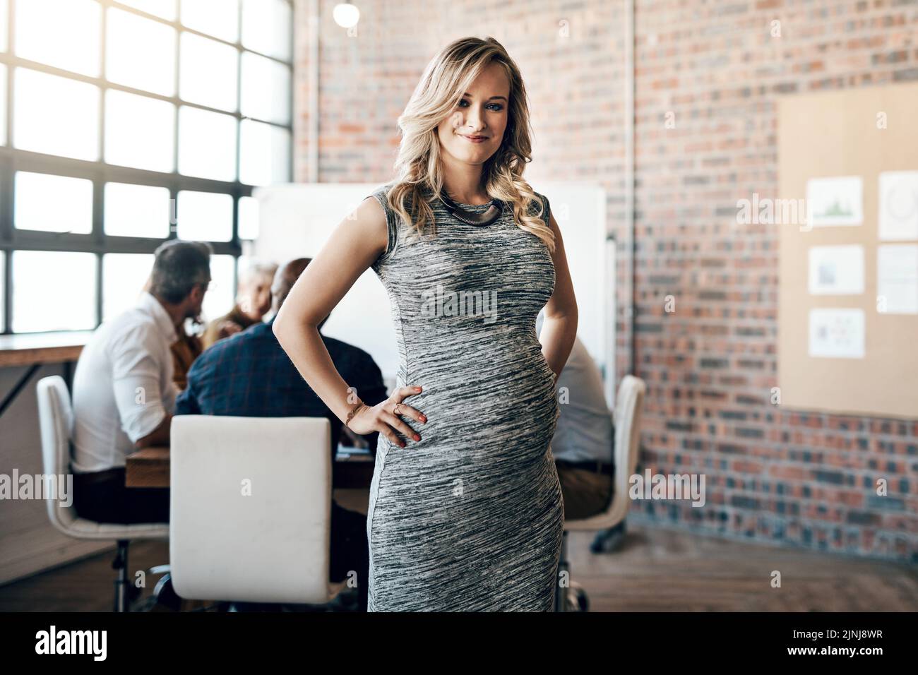 Modern and professional pregnant businesswoman smiling and standing in a leadership role, in a meeting with work colleagues. Ambitious mother working Stock Photo