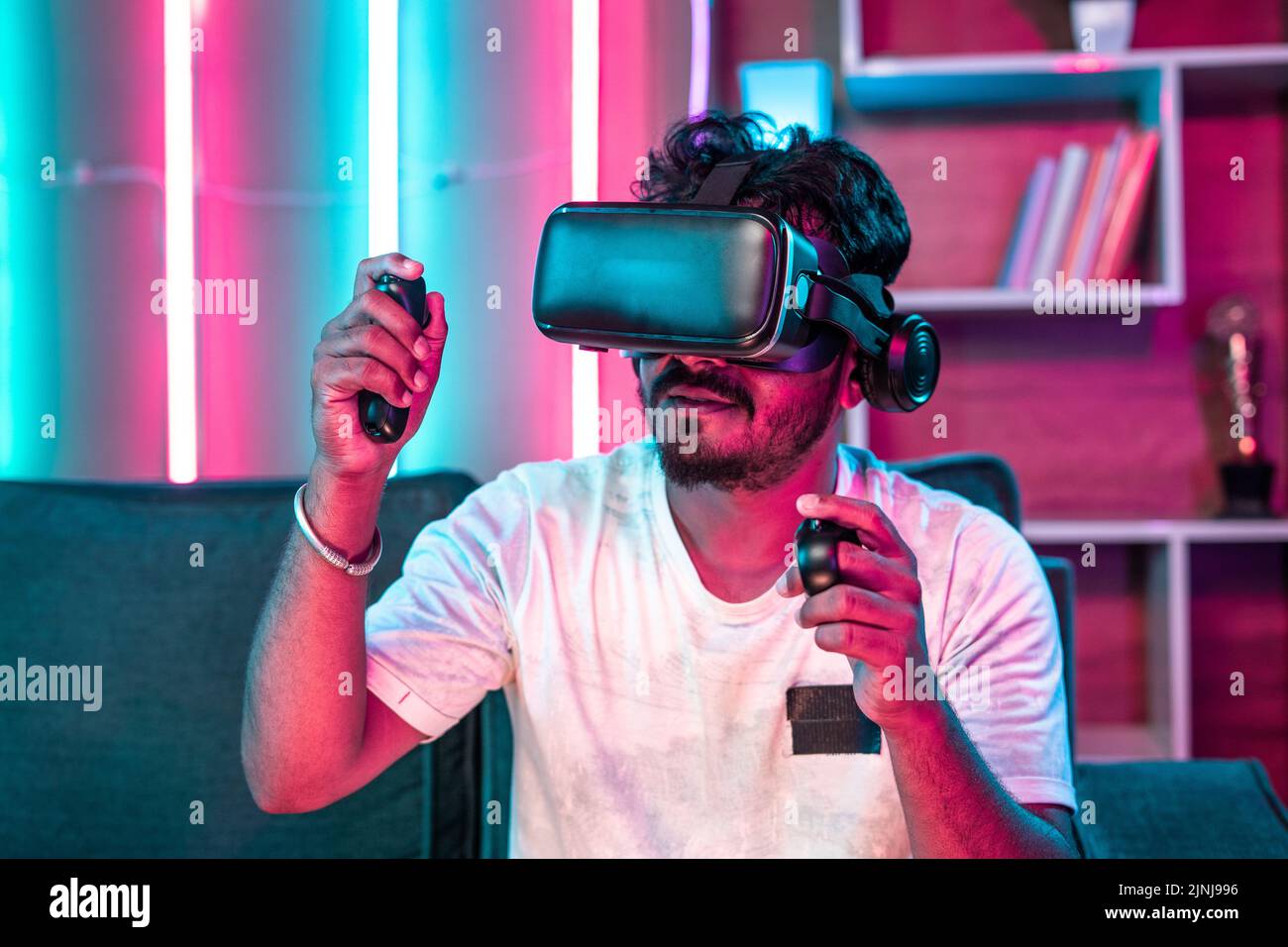 Focus on VR, Young gamer playing video game on virtual reality or VR headset using joystick at home - concept of metaverse, cyberspace and futuristic. Stock Photo