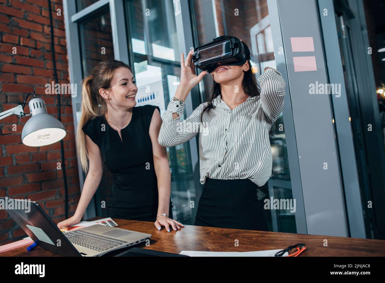 Two female application developers testing a new app designed for VR headset standing in modern office. Stock Photo