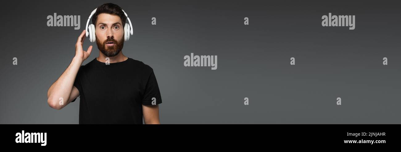 surprised man touching wireless headphones and listening music isolated on grey, banner,stock image Stock Photo