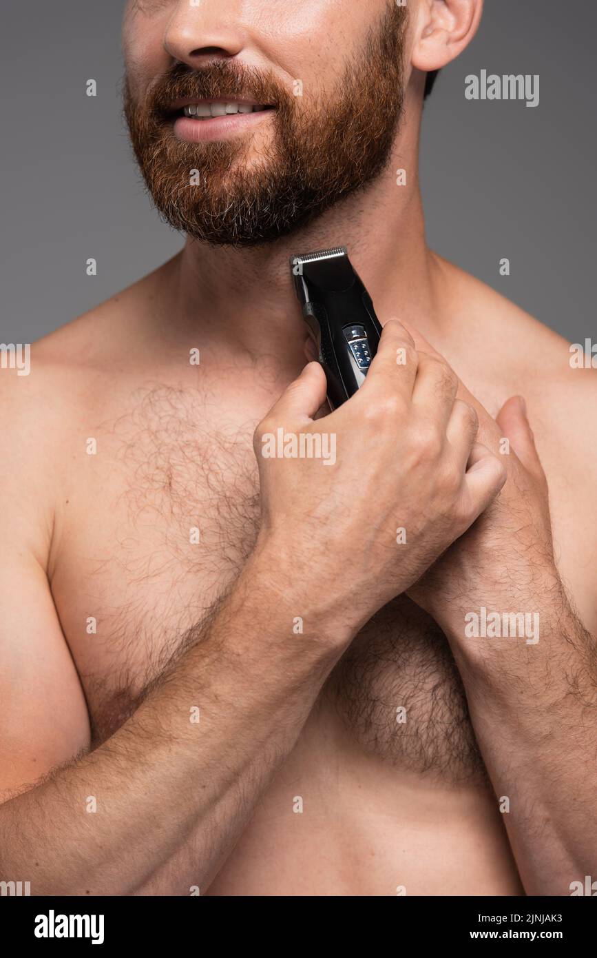 cropped view of shirtless man smiling and shaving beard with electric razor isolated on grey,stock image Stock Photo