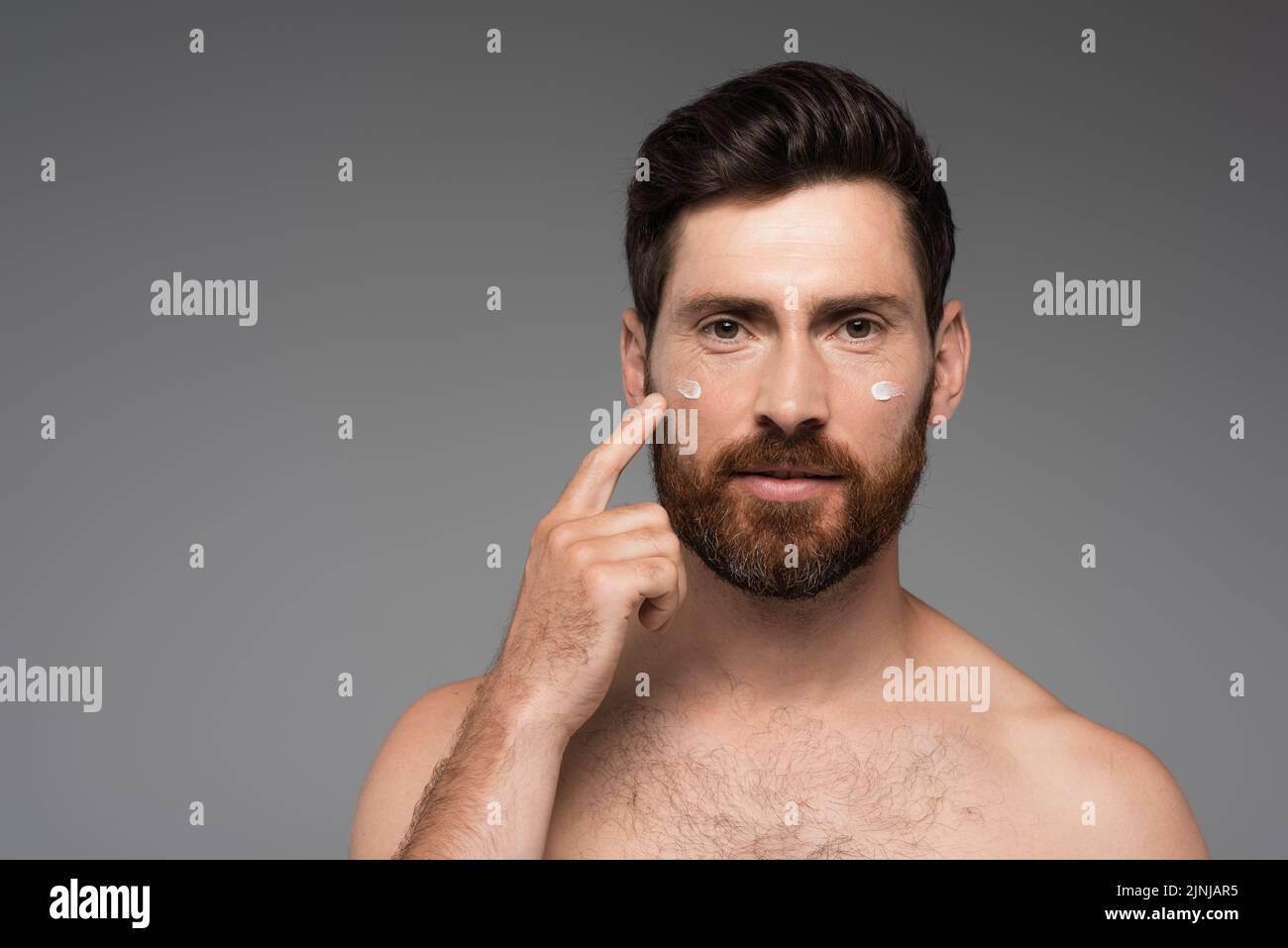 bearded man pointing with finger at cream on face isolated on grey,stock image Stock Photo