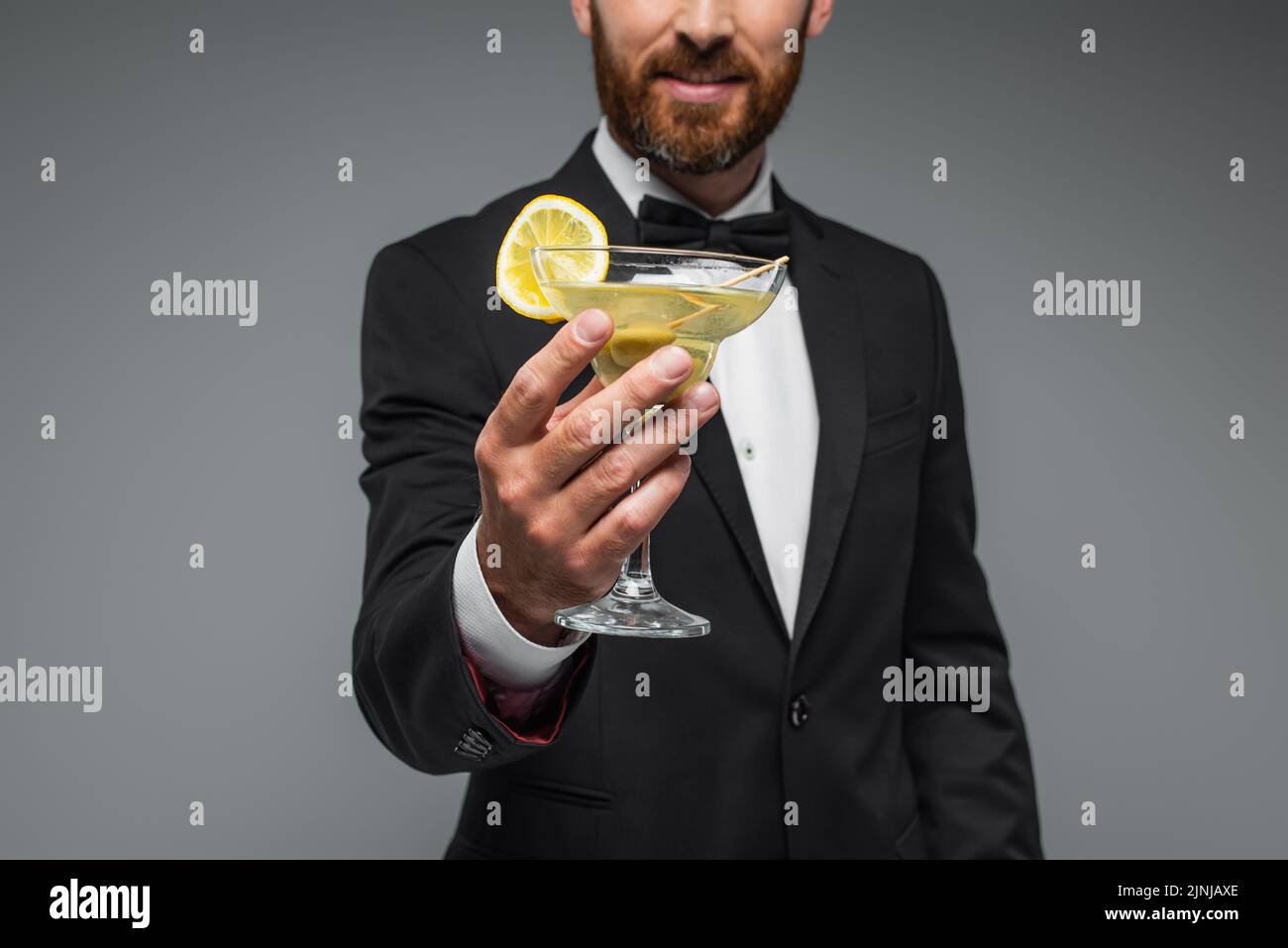 cropped view of cheerful and bearded man in suit with bow tie holding glass of cocktail isolated on grey,stock image Stock Photo
