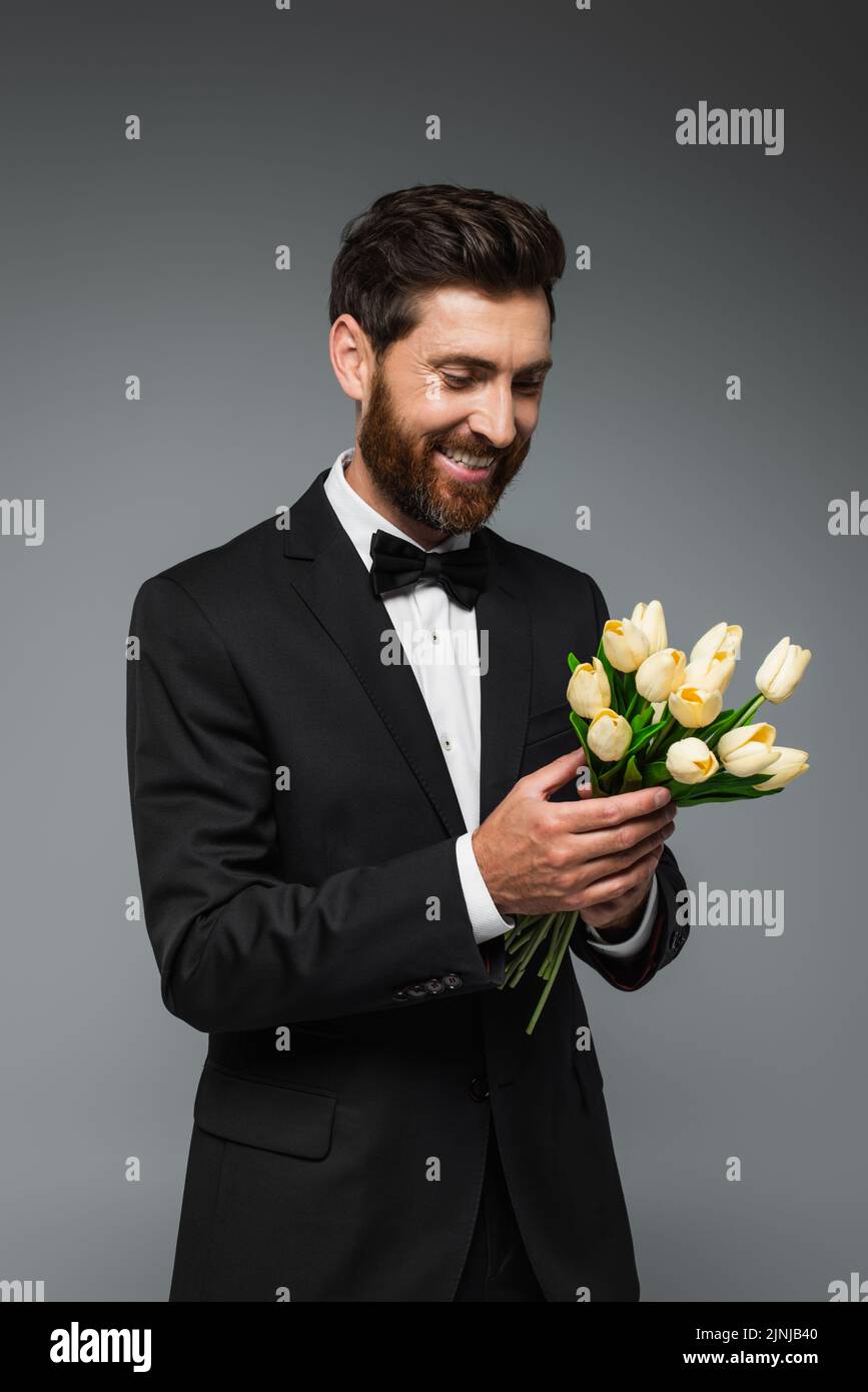 happy bearded man in elegant tuxedo with bow tie holding fresh tulips isolated on grey,stock image Stock Photo