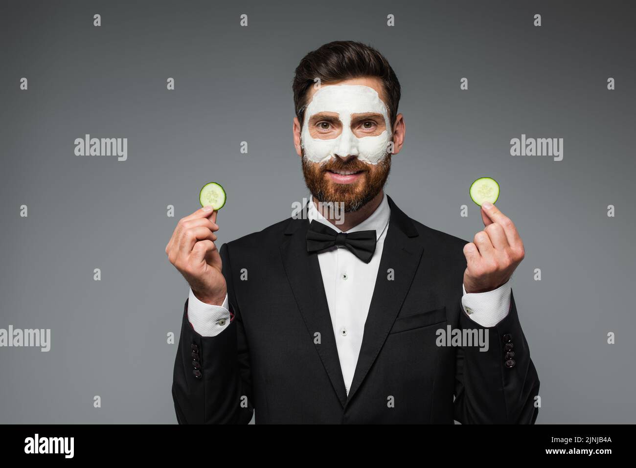 happy bearded man in suit with clay mask on face holding slices of cucumber isolated on grey,stock image Stock Photo