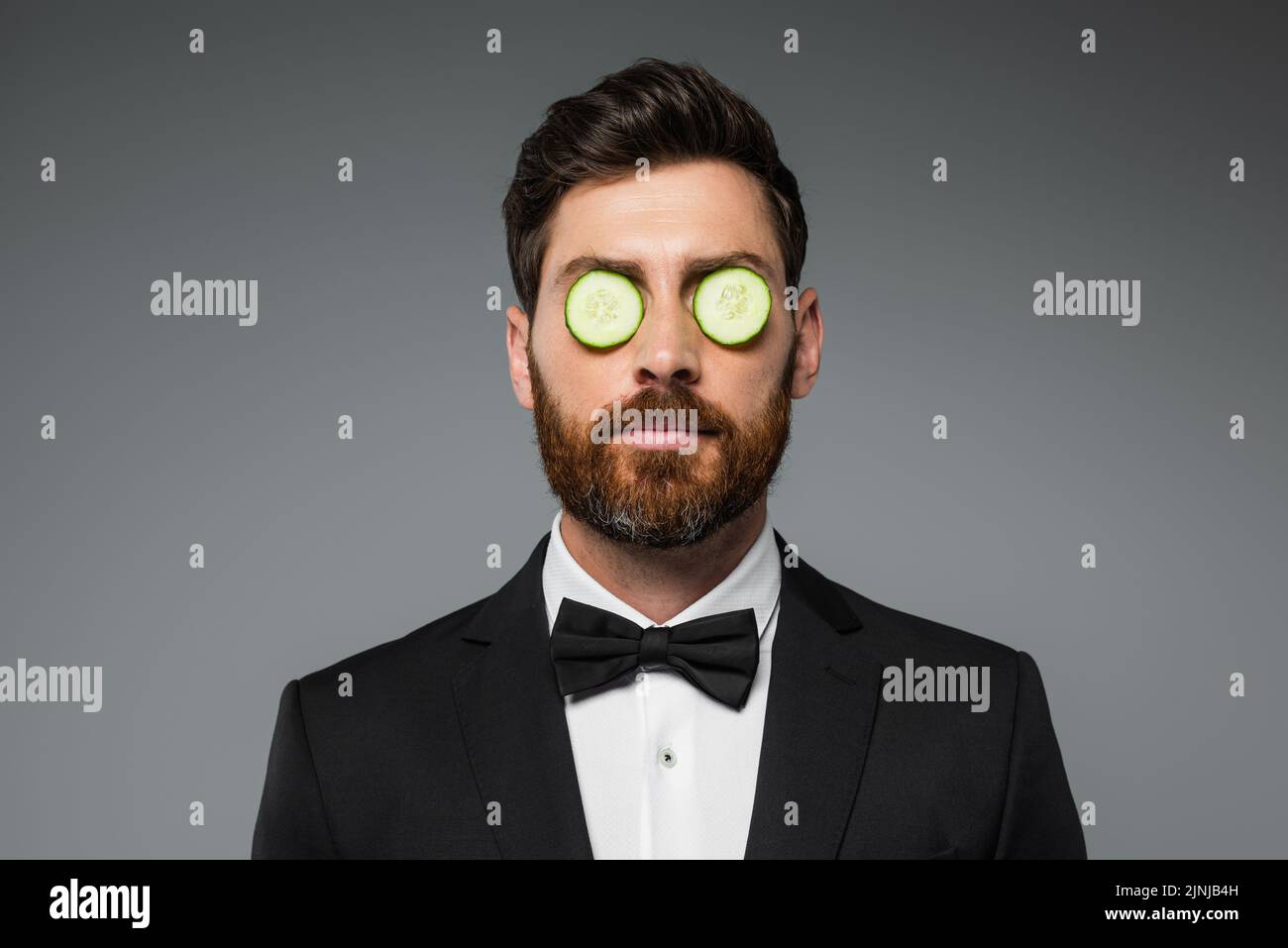 bearded man in tuxedo with fresh sliced cucumber on eyes isolated on grey,stock image Stock Photo