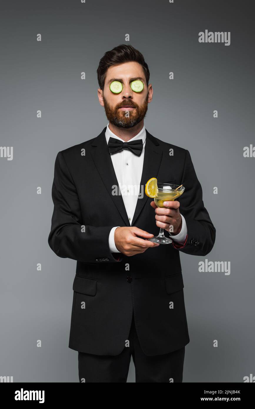 bearded man in tuxedo with fresh sliced cucumber on eyes holding glass of cocktail isolated on grey,stock image Stock Photo