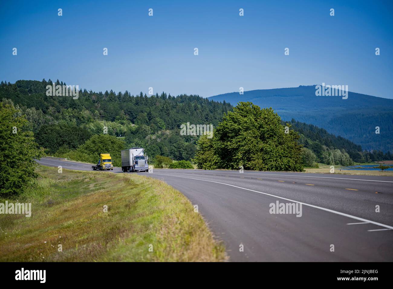Industrial blue and yellow tow big rig semi trucks transporting commercial cargo in dry van and flat bed semi trailers running on narrow highway road Stock Photo
