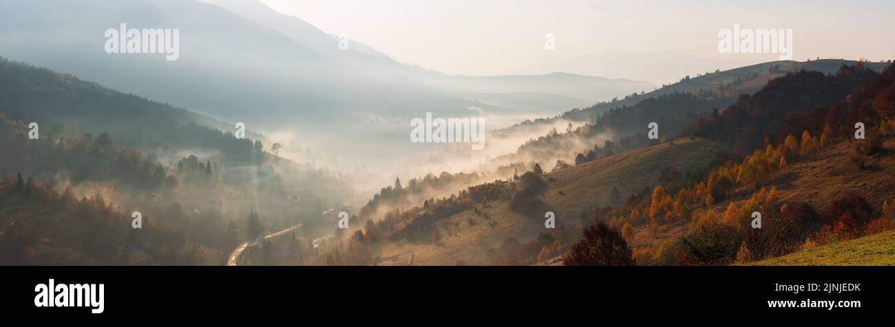countryside mountain landscape on a foggy morning. beautiful nature scenery with trees in colorful foliage on the hills and village in the distant val Stock Photo