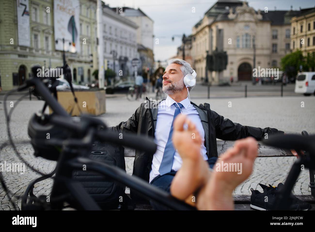 Businessman with bike sitting on bench, listening to music with feet up