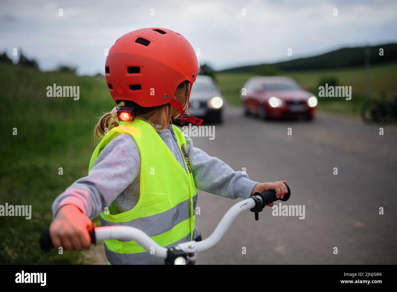 Rear view of little girl in reflective vest riding bike on road with cars behind her, road safety education concept. Stock Photo