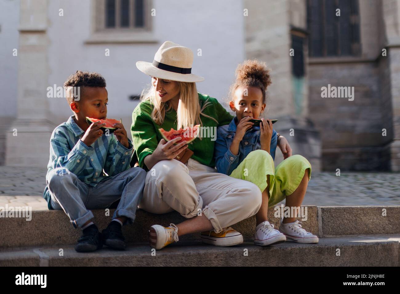 Multiracial kids with mother eating watermelon in street during hot sunny days, summer holiday travelling concept. Stock Photo