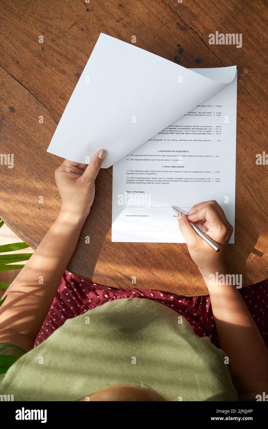 Female entrepreneur in casualwear sitting at wooden table and studying details of contract before signing it, directly above view Stock Photo